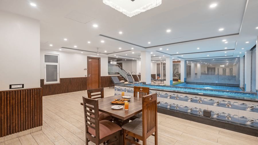 A bright, indoor swimming pool area at DLS Hotels On The Ganges. The pool is a vibrant blue, and a row of dark sun loungers with yellow and white striped towels are placed on a light-colored tiled floor. The walls are white with blue mosaic tile accents,