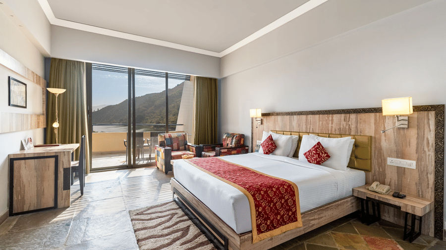 A bed with a runner on it and a large window on one side with a lake view at Via Lakhela Resort & Spa, Kumbhalgarh