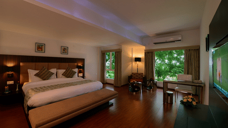 duplicate of a queen bed room with air-conditioning, wall-mounted TV, workstation, a seating area with a sofa, and two separate glass windows showcasing the exterior lush greenery at Top3 Lords Resort Bhavnagar