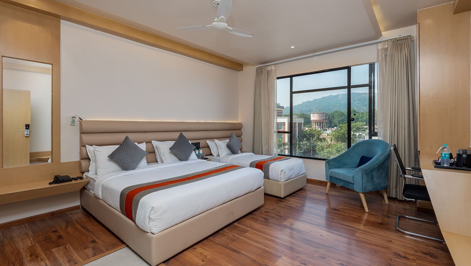 An image of a room with a king size bed and a single bed along with a chair, and a glass window overlooking the city view - DLS Hotel Arihant, Haridwar