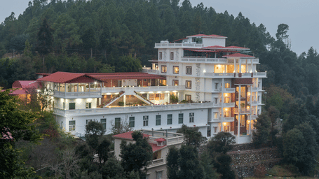 Exterior of the Tehri Club Resort, Chamba, by DLS hotels, featuring the large hotel building against a forested hill under a dark cloudy sky.