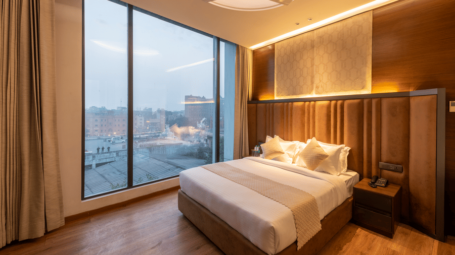 A bedroom with a king size bed, side tables, ceiling fan, lamps, and a large window with curtains at DLS Hotels Amritsar.