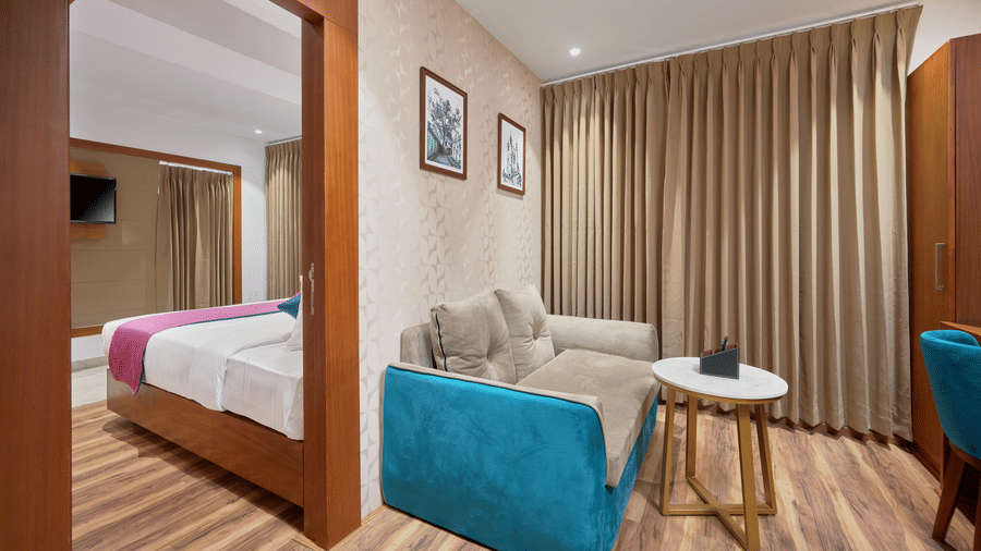View of a suite at QWhite Crescent Luxury Hotel by Dimora Kochi with a teal sofa, round side table, wooden flooring, and bedroom beyond.