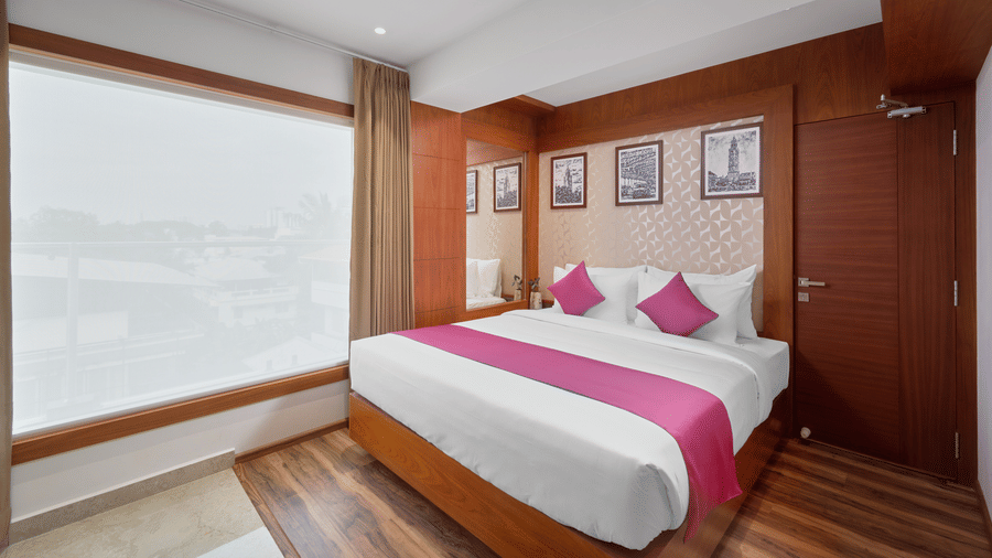 View of a room at QWhite Crescent Luxury Hotel by Dimora Kochi with a king bed, pink accent runner, wooden panels, and a large window.