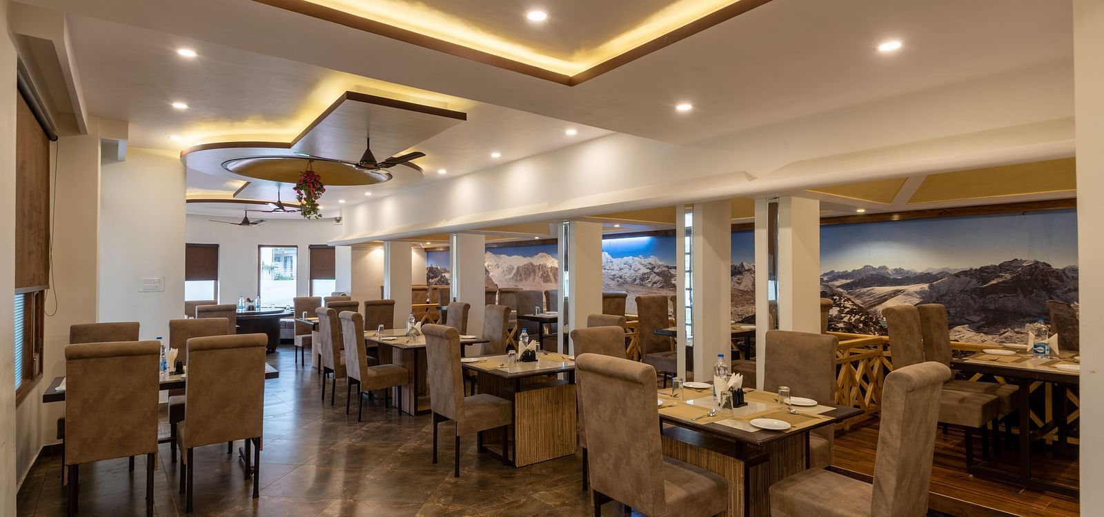 A spacious restaurant in Bhagsu with a modern design, featuring large mountain murals on the walls and rows of tables and chairs – DLS Meghavan Resort.