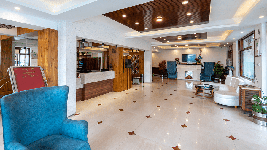 A hotel reception area with a blue armchair, a welcome sign, a reception desk, and white seating - DLS Meghavan Resort, Dharamshala