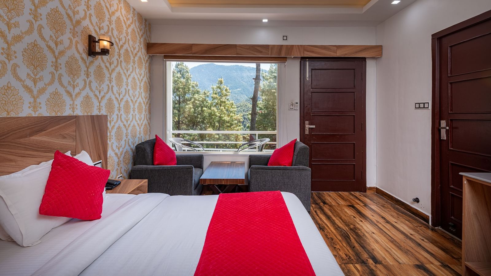  Cosy hotel room with a bed featuring red accents, stylish wallpaper, wooden flooring, seating area by a large window, and views of trees and distant hills - DLS Nature Trinket Resort, Dalhousie.