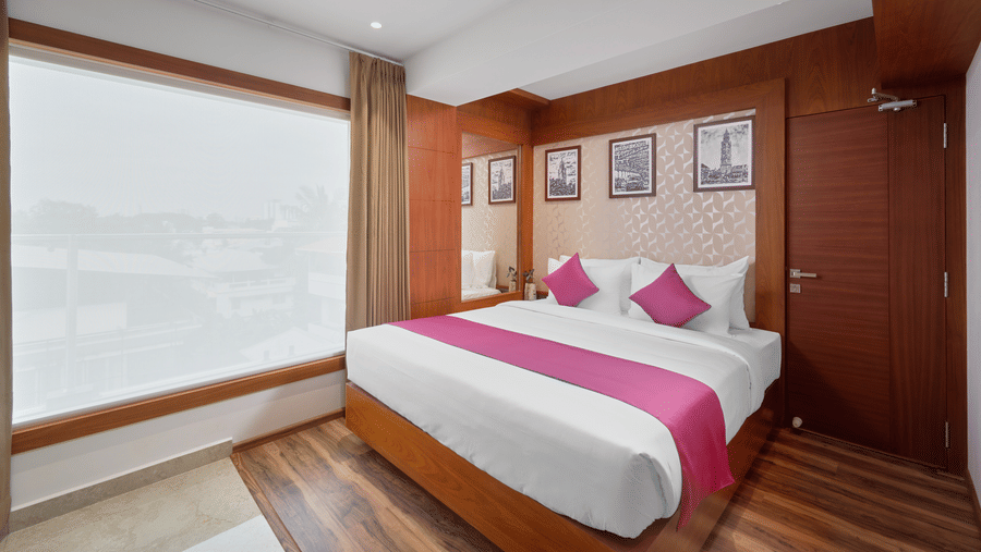 View of a room at QWhite Crescent Luxury Hotel by Dimora Kochi with a king bed, pink accent runner, wooden panels, and a large window.