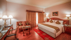 A warmly lit hotel room with a king sized bed, classic wooden decor, work desk, and cosy seating area – Raj Park Hotel, Chennai