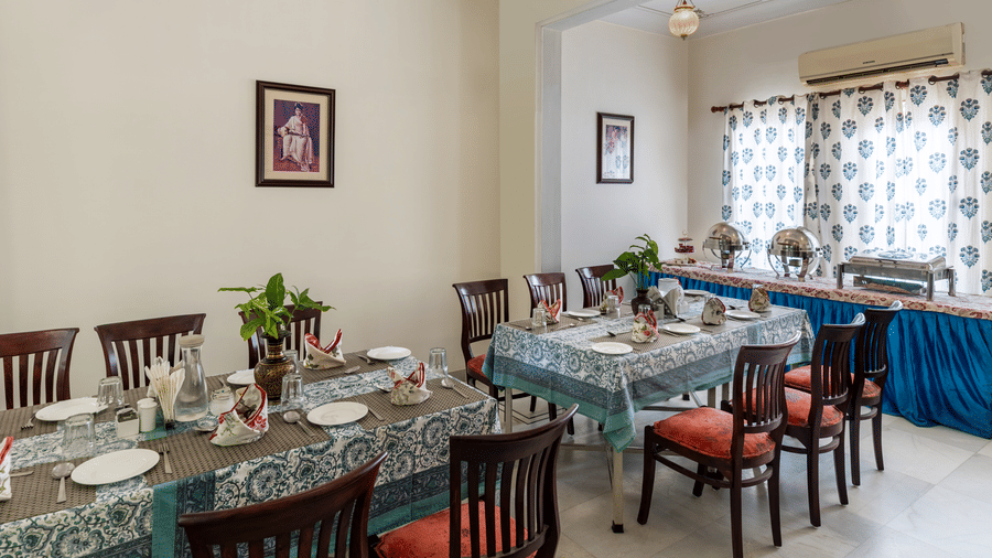 A dining area with 2 tables with patterned tablecloths, chairs, and framed pictures at Akshay Niwas Boutique Hotel by Amantra, Udaipur.