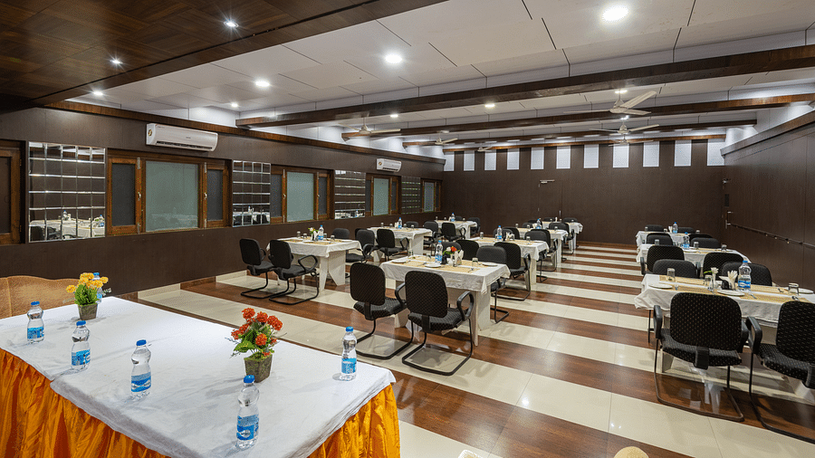 A conference hall in Bhagsu with arranged tables, chairs, and decorative flowers. The room features wooden panelling, bright lights, and large windows - DLS Meghavan Resort, Dharamshala