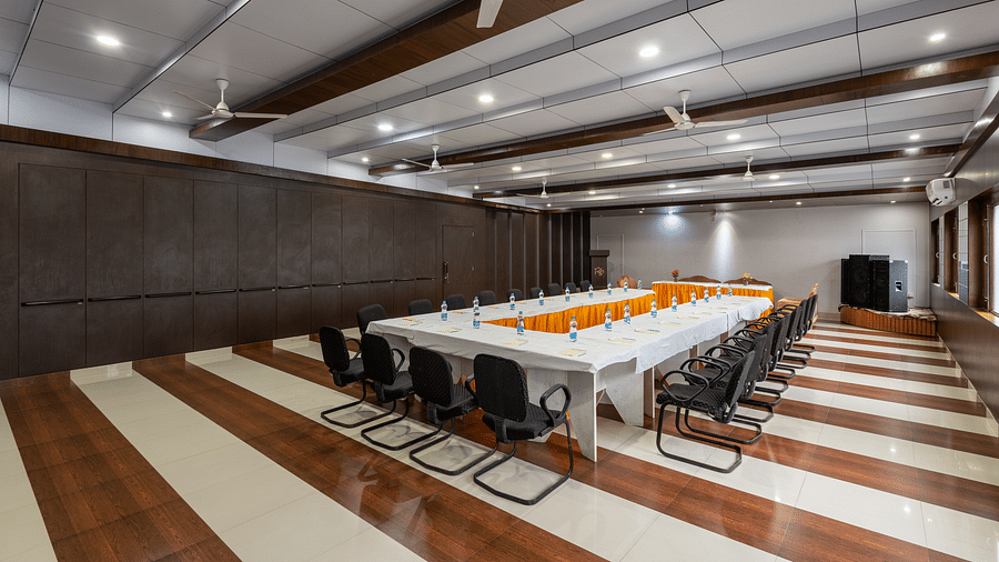 A conference room with a long table, chairs, and decorative flowers. The room features wooden panelling, bright lighting, ceiling fans, and windows along the walls - DLS Meghavan Resort, Dharamshala