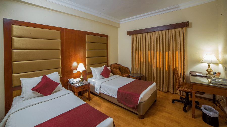 An overview of the twin beds in a inside Deluxe Rooms at Raj Park Hotel, Chennai, featuring a table, a lamp, a study table and a chair. This hotel is one of the best places to stay in Chennai with it's connectivity and convenience.