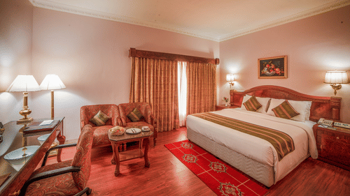 A warmly lit hotel room with a king sized bed, classic wooden decor, work desk, and cosy seating area – Raj Park Hotel, Chennai