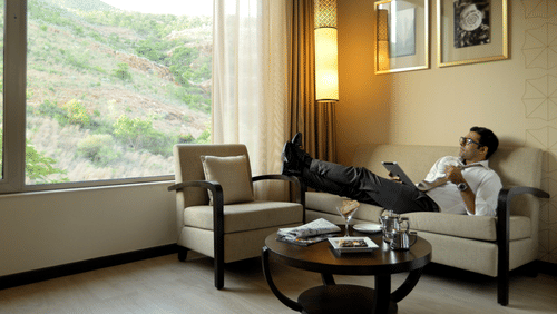 Guest relaxing in the Deluxe Room at Marasa Sarovar Premiere, Tirupati, featuring a comfortable seating area, large picture window with hillside views, and warm contemporary decor.