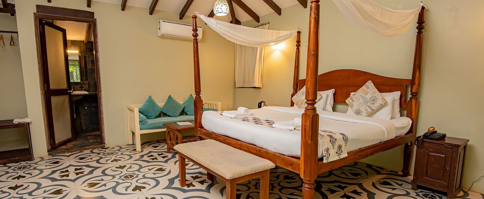 Spacious room with a large bed, wooden seating at the foot, bedside table, and a doorway in the background at De Soul Santé, Morjim.