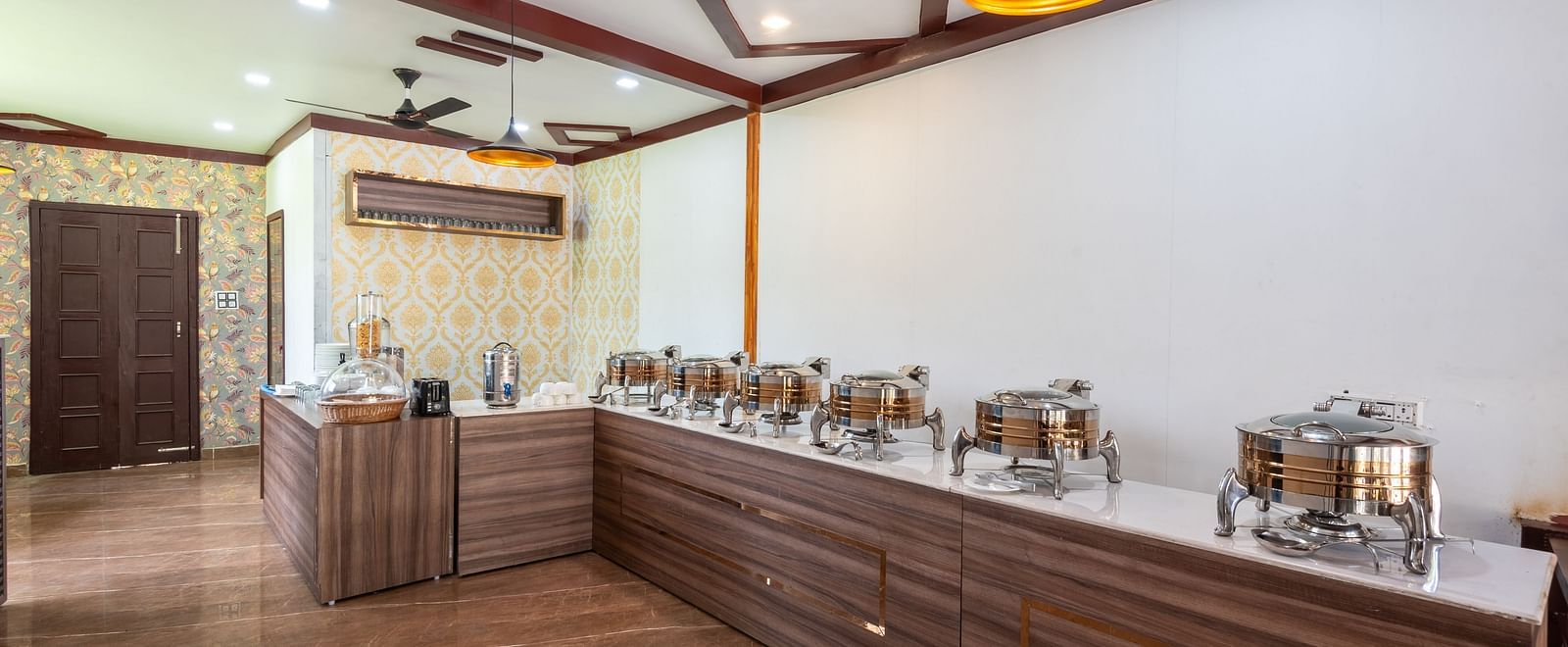 Buffet area in a restaurant featuring a long wooden counter with chafing dishes, warm lighting, and decorative wall accents - DLS Nature Trinket Resort, Dalhousie.