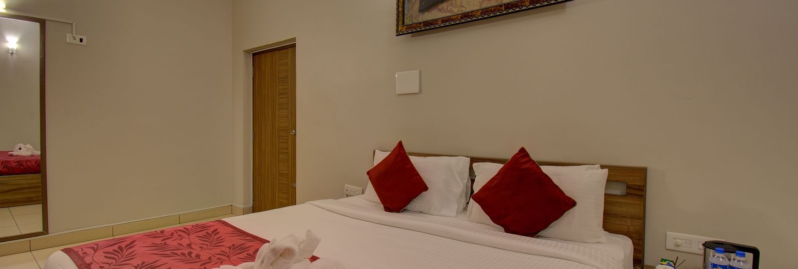 A swan made with of towel on a huge bed, a side table, mirror, wooden door and an wall art | Superior Room | Woodstock Resorts, Coorg
