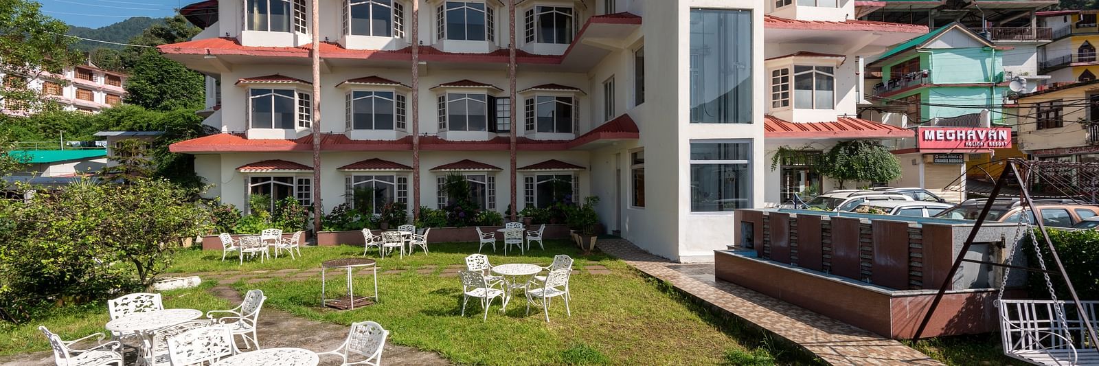 A resort building with a garden, white outdoor tables and chairs, and a swing, set against a hilly backdrop - DLS Meghavan Resort, Dharamshala