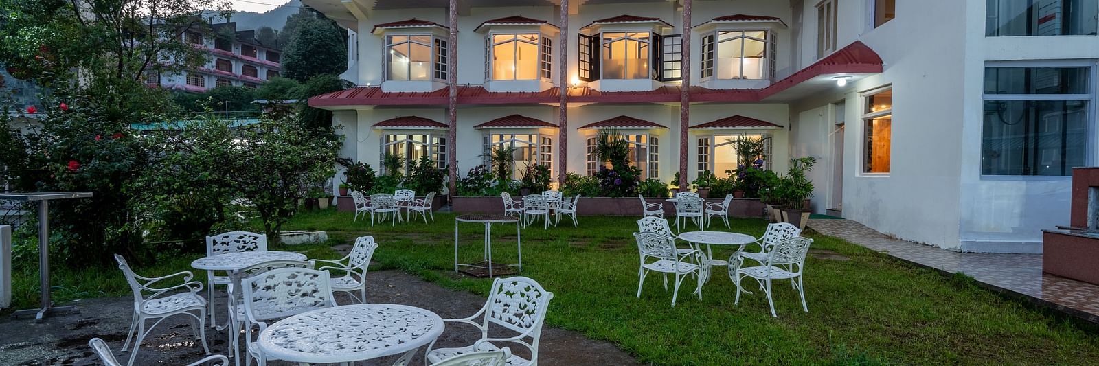A resort garden with white outdoor tables and chairs, set against a building with lit windows. The area is surrounded by greenery and plants - DLS Meghavan Resort, Dharamshala