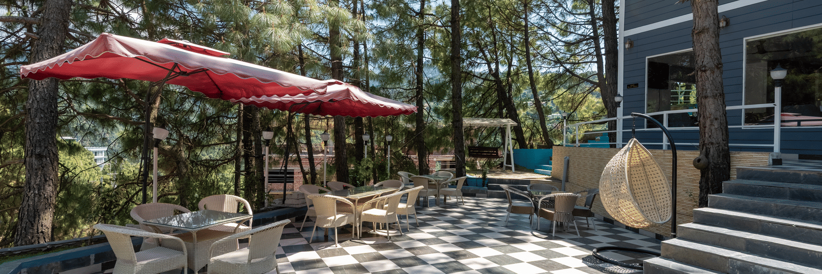 Outdoor patio with black-and-white checkered flooring, red umbrellas, beige seating, and a staircase leading to a grey building, surrounded by tall pine trees - DLS Nature Trinket Resort, Dalhousie.