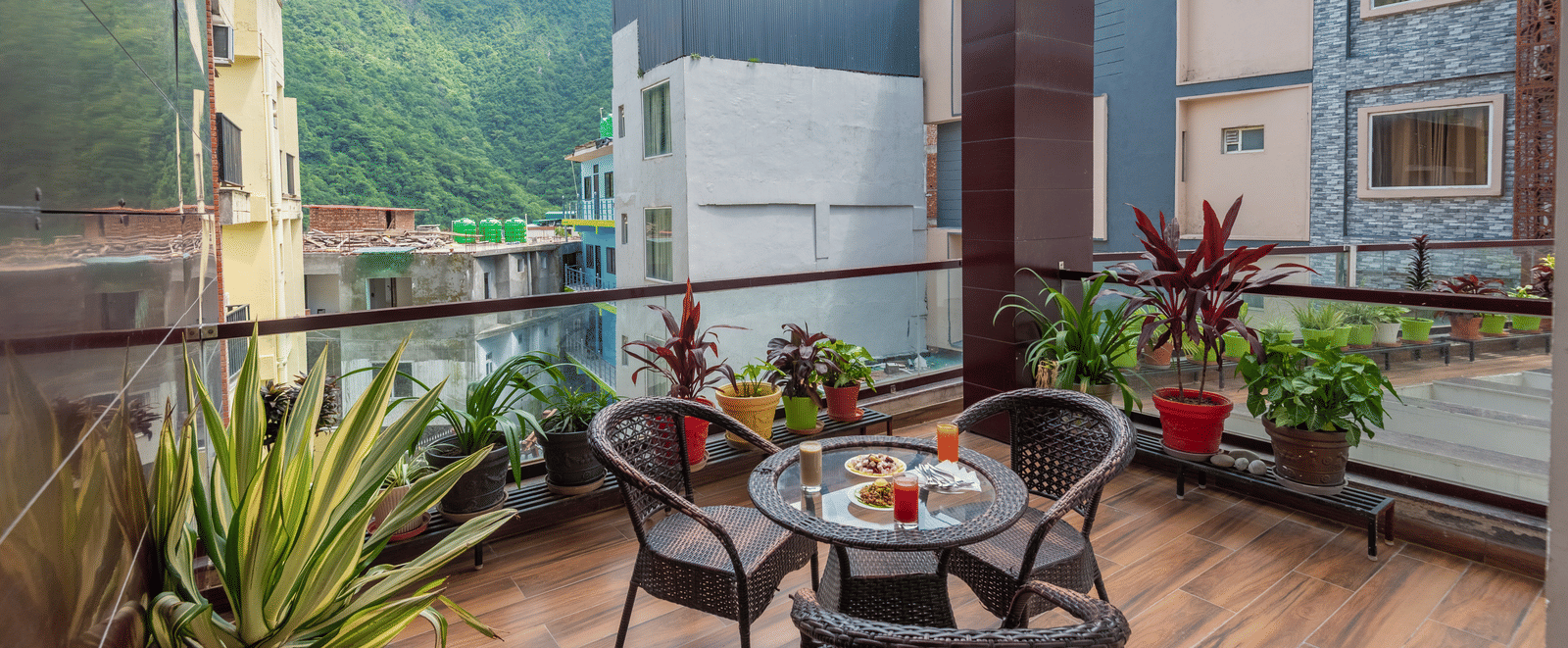 A wide view of a balcony at DLS Hotels On The Ganges, offering a scenic vista of green hills and surrounding buildings under a blue sky with white clouds. A small wicker dining set with food and drinks sits on the wood-like tiled floor, and various potte