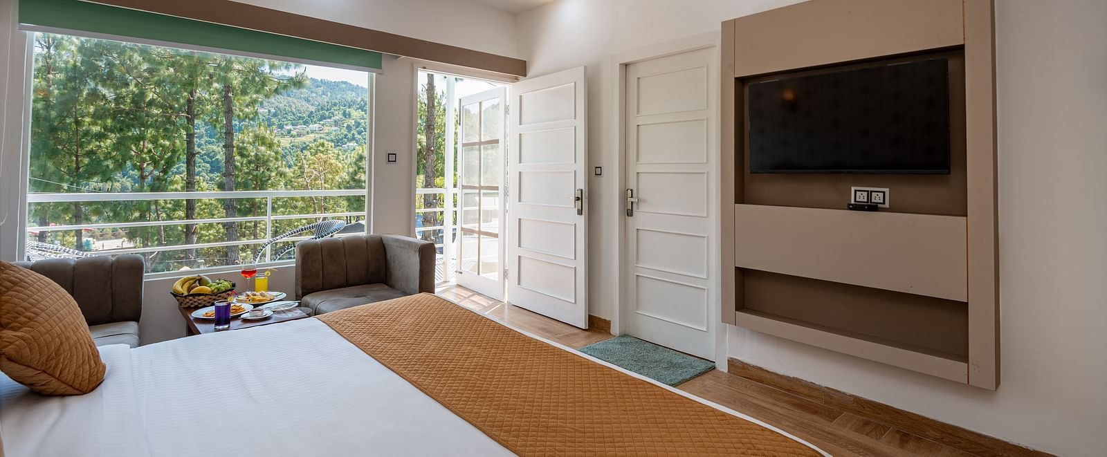 A bright hotel room with a double bed, TV, and seating area, opening onto a balcony with scenic hill views - DLS Nature Trinket Resort, Dalhousie.