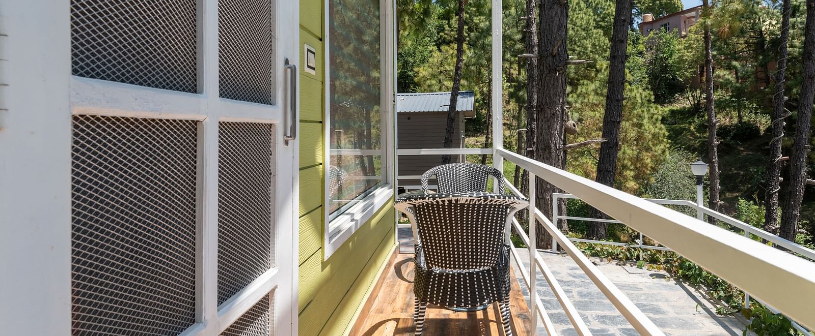  Narrow wooden balcony outside a green cottage with white railings, featuring a small table and chairs, overlooking a peaceful forested area with tall trees - DLS Nature Trinket Resort, Dalhousie.