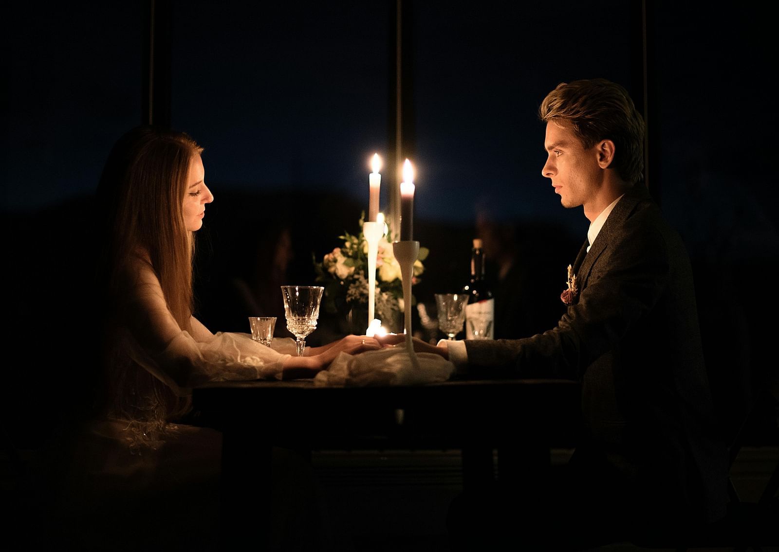 A romantic couple sharing an intimate candlelit dinner at night, holding hands across the table with wine glasses and flowers nearby.