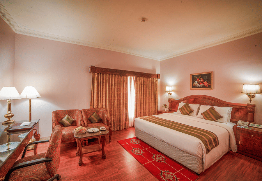 A bedroom at Raj Park Hotel, Chennai, featuring a king size bed, sofa, bedside tables with lamps, and a work desk with chair.