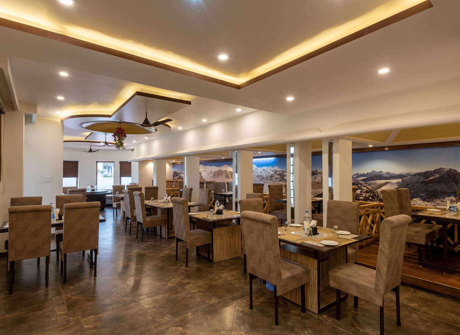 A spacious restaurant in Bhagsu with a modern design, featuring large mountain murals on the walls and rows of tables and chairs – DLS Meghavan Resort.