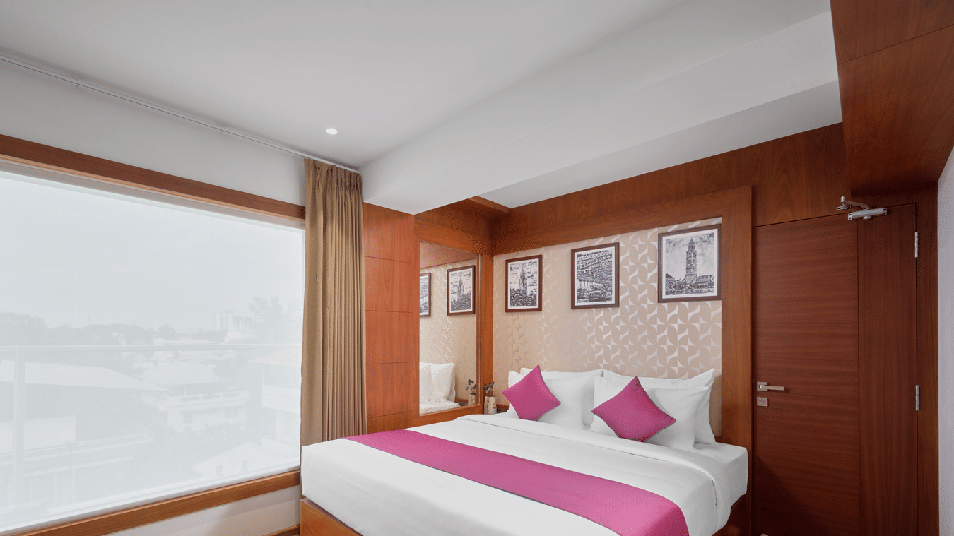 View of a room at QWhite Crescent Luxury Hotel by Dimora Kochi with a king bed, pink accent runner, wooden panels, and a large window.