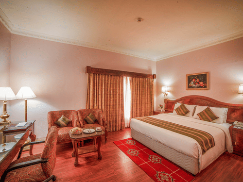 A warmly lit bedroom at Raj Park Hotel - Chennai featuring a plush bed by the window and a sitting area at its foot.