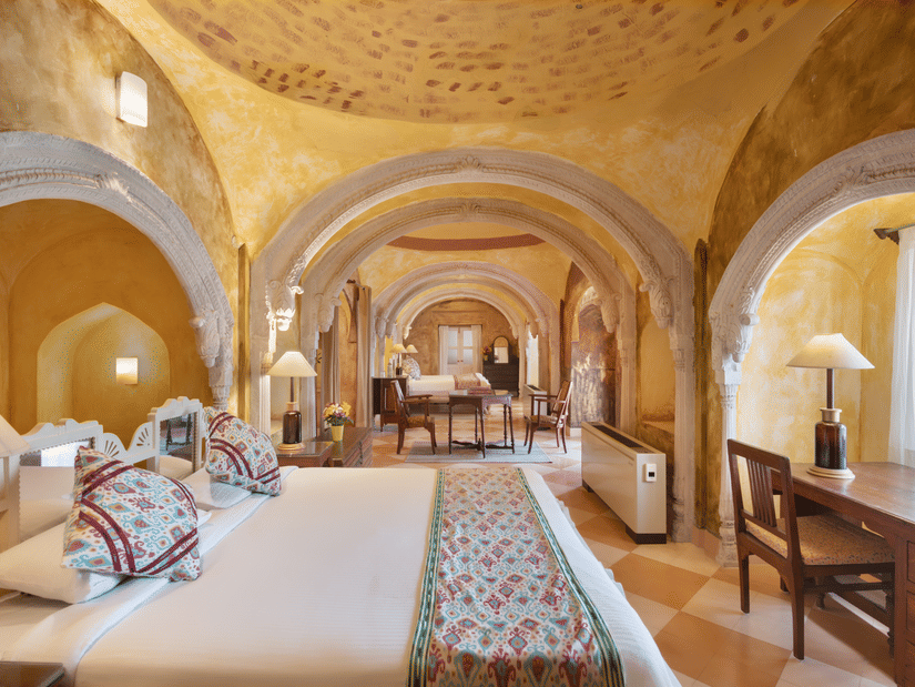 A room with arched doorways, a patterned ceiling, and a view into a dining area, featuring a bed with decorative pillows and a wooden desk, showcasing the fort-palace's unique design - Tijara Fort-Palace - 19th Century, Alwar.