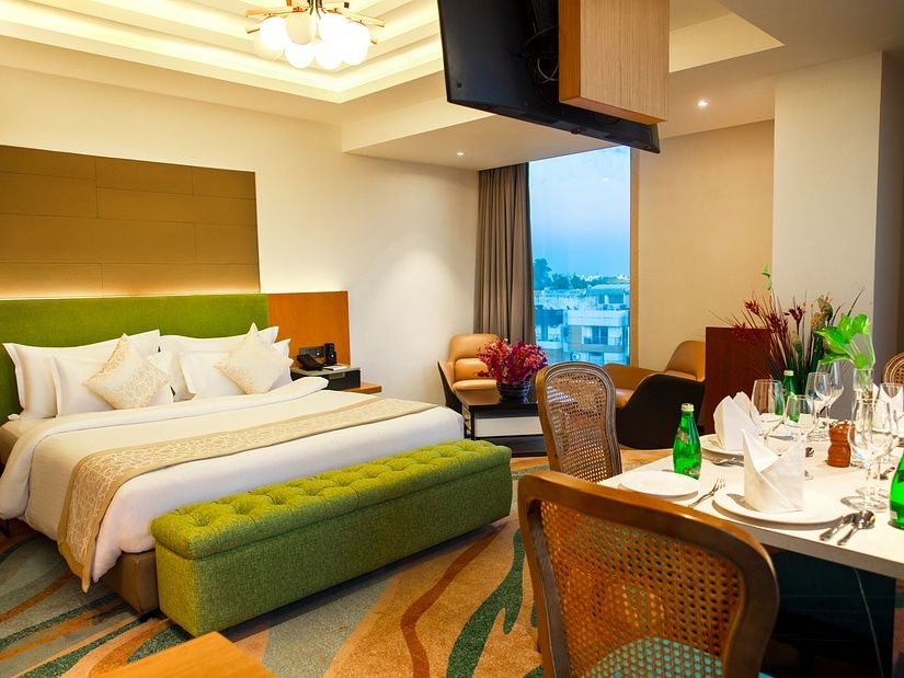 Luxurious hotel room with a king-size bed, modern decor, city view through large window, and an elegant dining setup with bottled water at The Regnant .