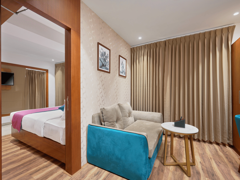 View of a suite at QWhite Crescent Luxury Hotel by Dimora Kochi with a teal sofa, round side table, wooden flooring, and bedroom beyond.