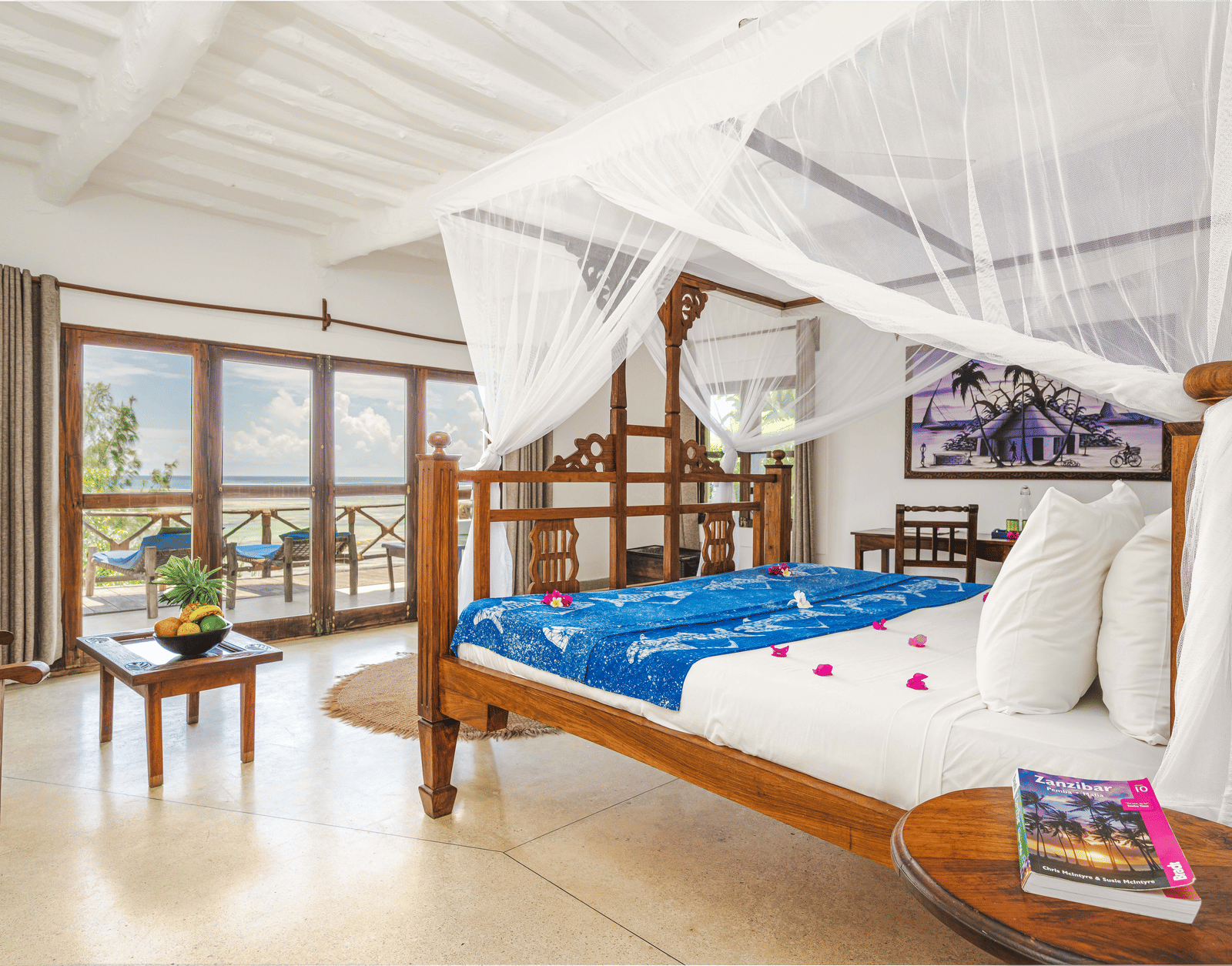 View of a room showcasing a large bed with mosquito nets and decorative pillows, a small table, and artistic wall hangings in Sea View Family Cottages - Shooting Star Boutique Hotel.