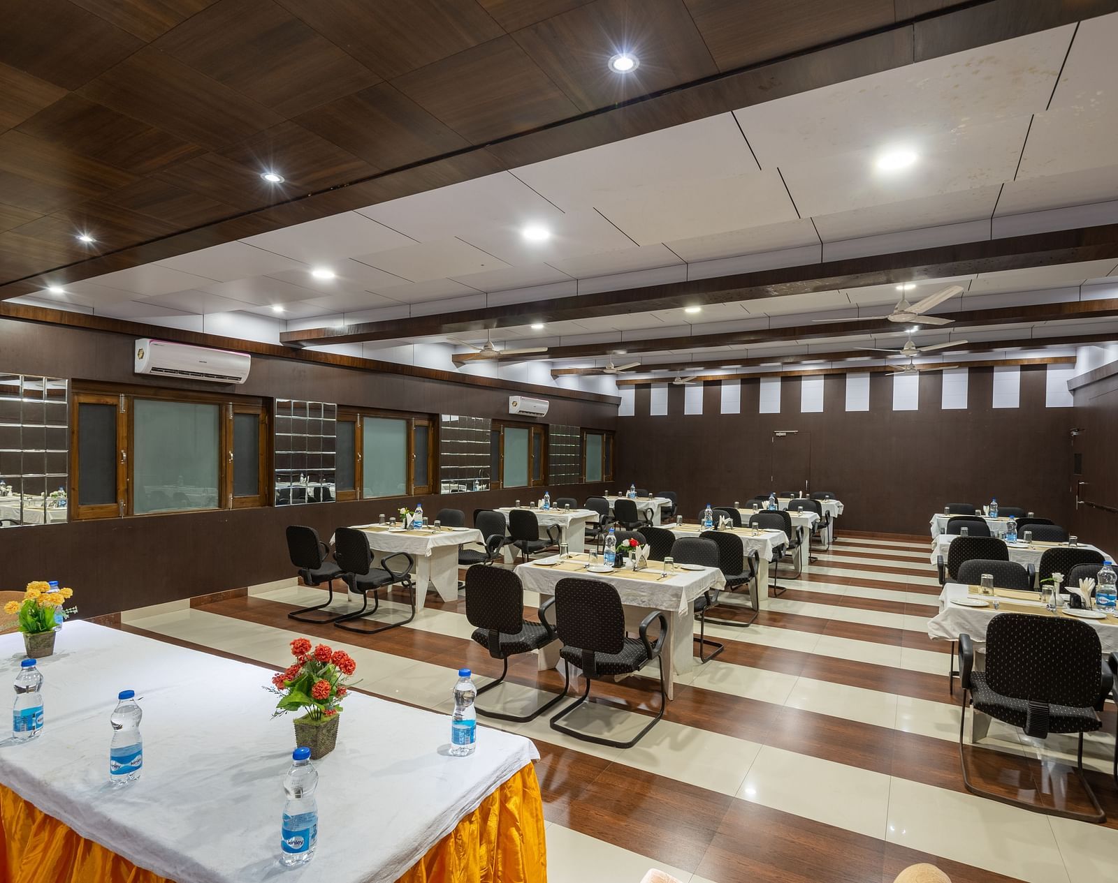 A conference hall in Bhagsu with arranged tables, chairs, and decorative flowers. The room features wooden panelling, bright lights, and large windows - DLS Meghavan Resort, Dharamshala