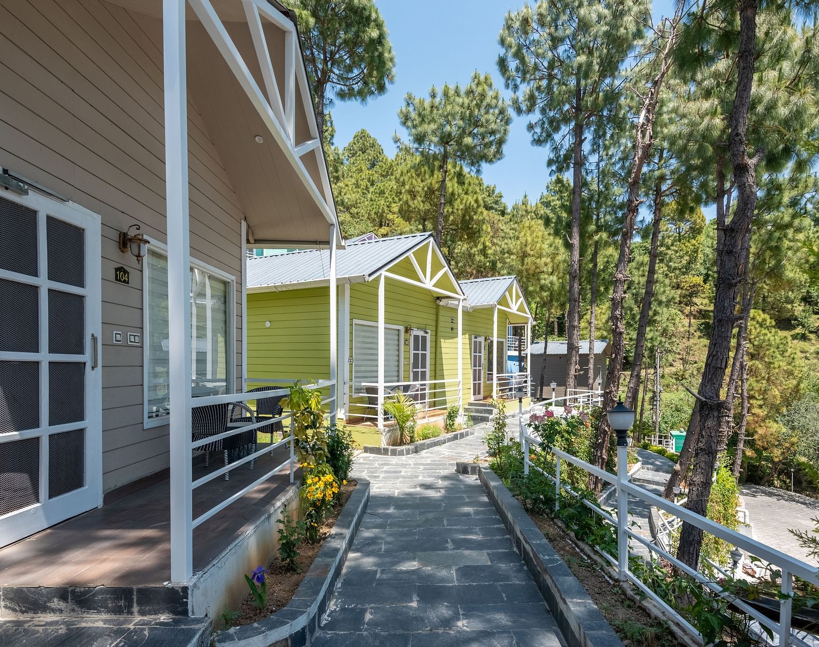 A paved pathway stretches between rows of cabins with light-coloured walls and dark window frames, nestled among tall green trees - DLS Nature Trinket Resort, Dalhousie.