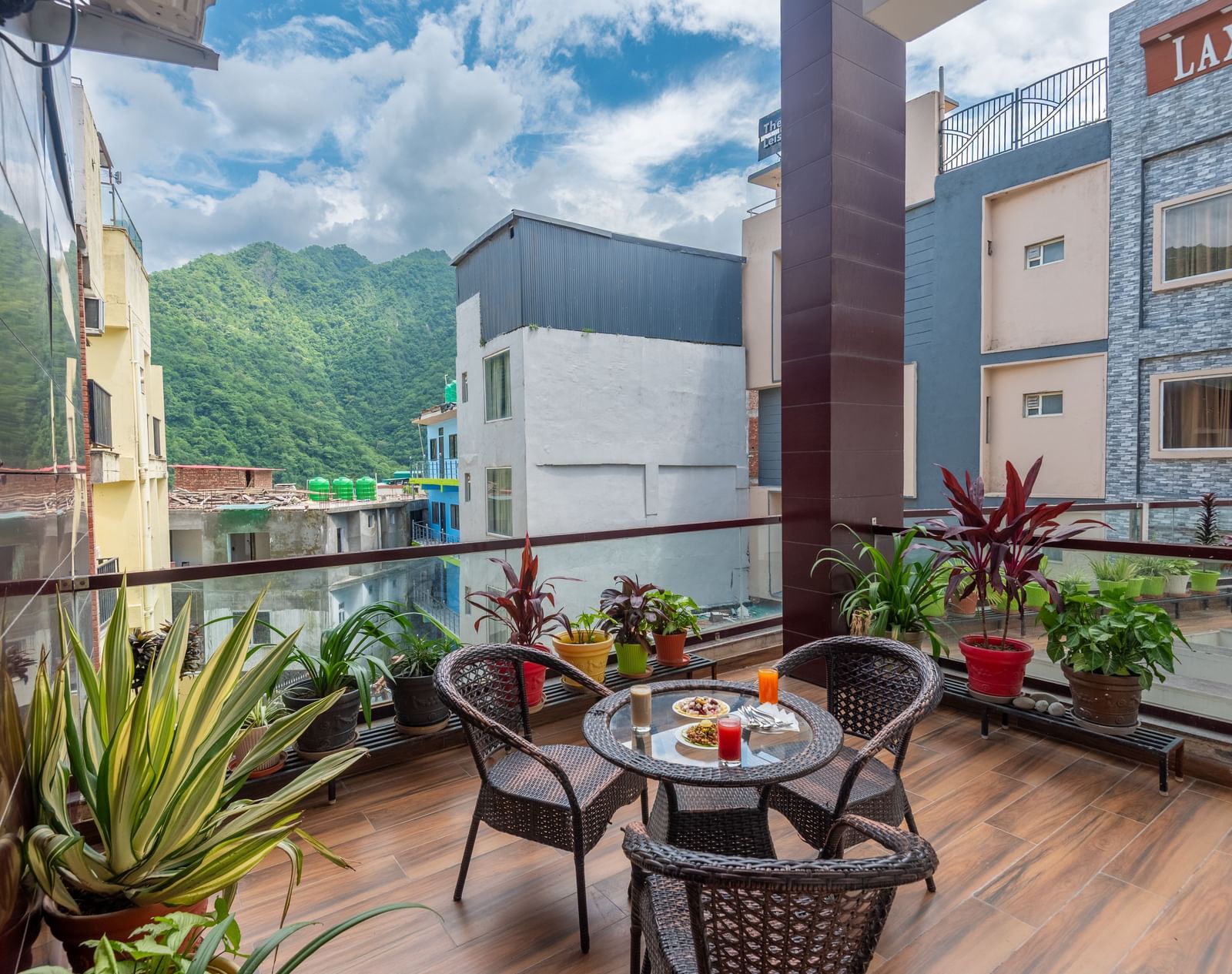 A wide view of a balcony at DLS Hotels On The Ganges, offering a scenic vista of green hills and surrounding buildings under a blue sky with white clouds. A small wicker dining set with food and drinks sits on the wood-like tiled floor, and various potte
