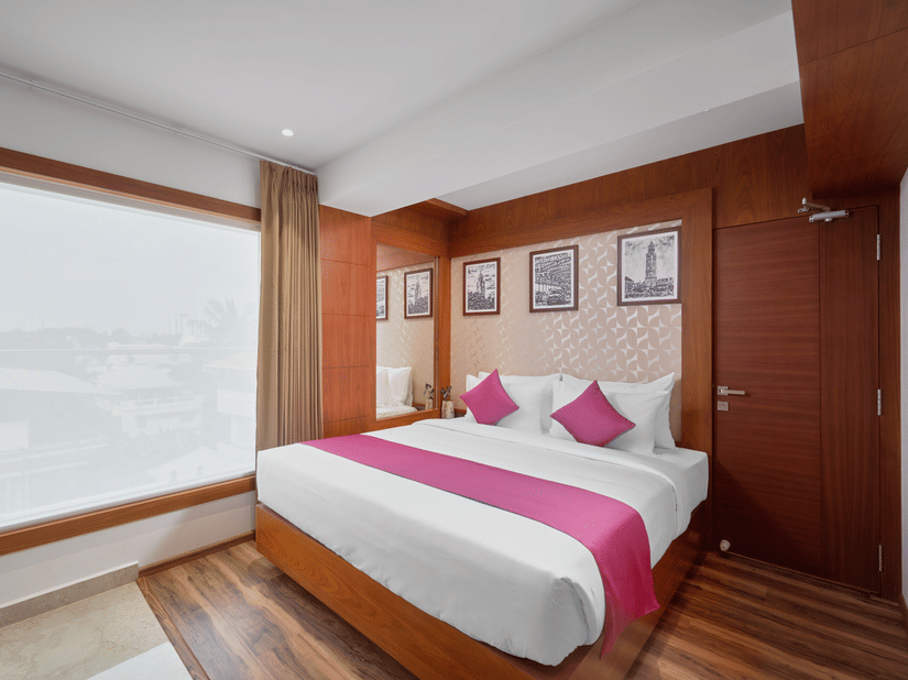 View of a room at QWhite Crescent Luxury Hotel by Dimora Kochi with a king bed, pink accent runner, wooden panels, and a large window.