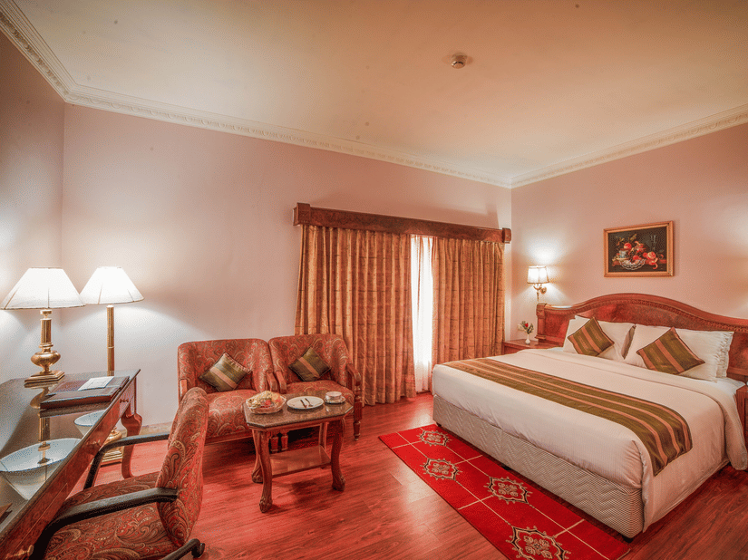 A bedroom at Raj Park Hotel, Chennai, featuring a king size bed, sofa, bedside tables with lamps, and a work desk with chair.