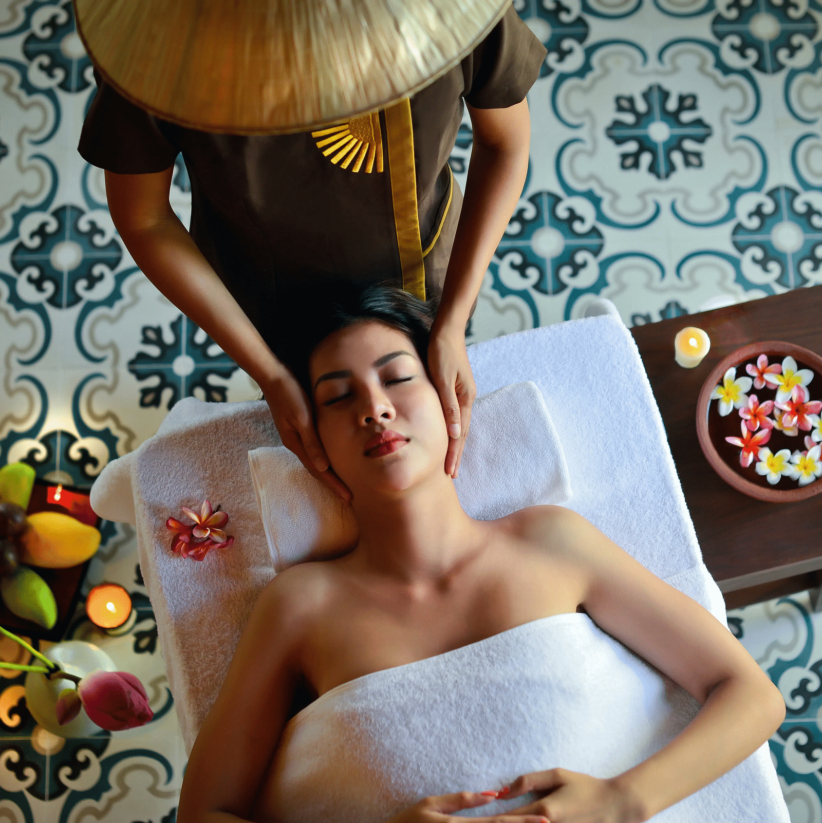 A woman wrapped in a white towel receives a relaxing head massage in a serene spa setting, surrounded by flowers and candles.