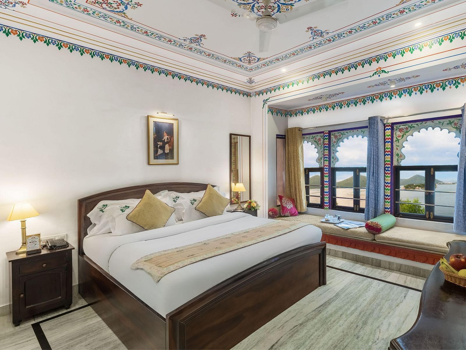  A spacious bed with a wooden bedside table and a label, alongside bay window seating with curtains pulled to the side at Panna Vilas Palace