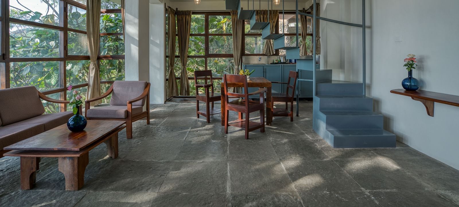 Spacious living area with a blue staircase, wooden furniture, and expansive windows providing a view of the lush greenery outside. | Mango Hill, Yercaud