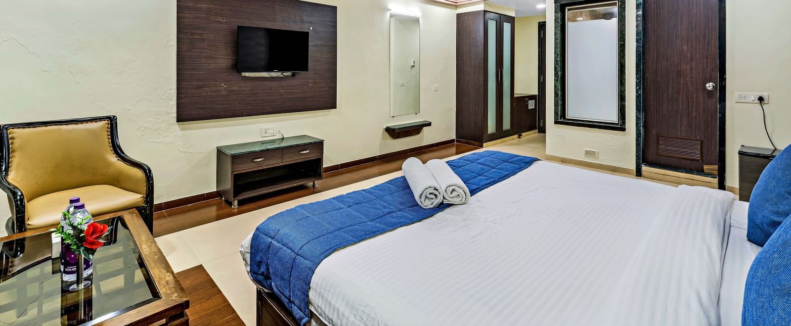 A wide view of a suite in Krushnai Resort featuring a double bed with blue runner, a wall mounted TV, modern furnishings and a lounge chair.