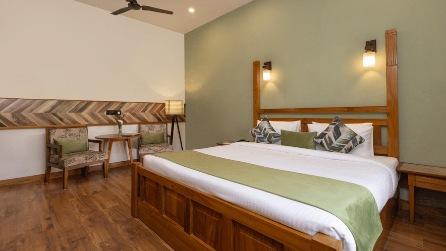 A hotel bedroom with a large wooden bed frame, a green bed runner, a small couch, and a large window at The Golden Tusk, Jim Corbett