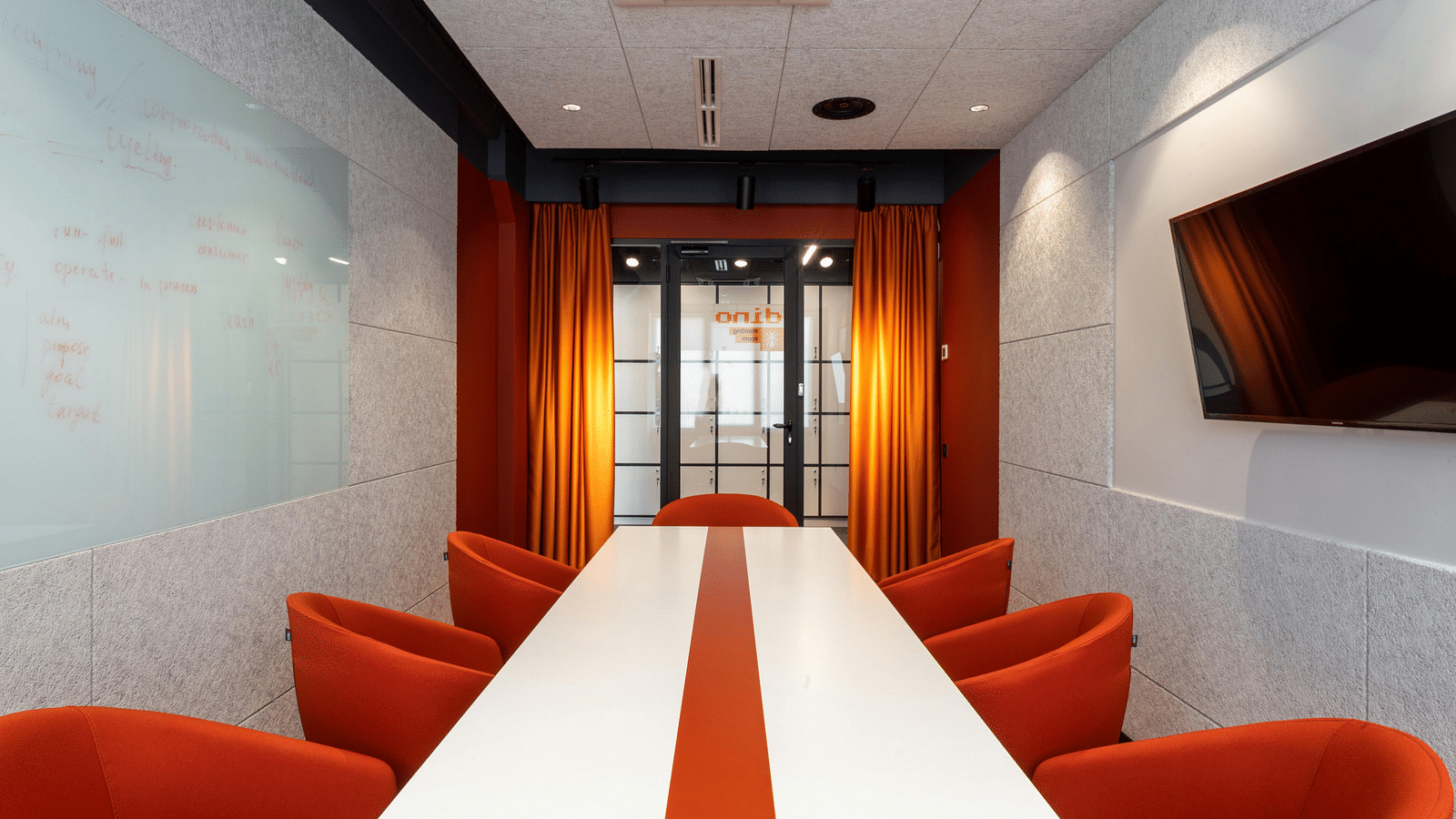 A conference room with a long whiteboard, a white and red striped table, orange chairs and red curtains partially concealing a glass door