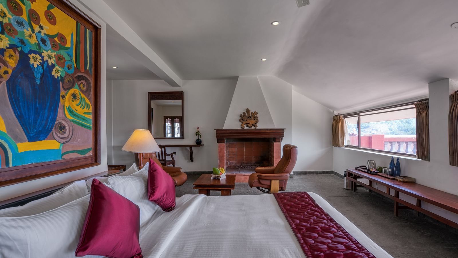 Spacious hotel room with a cozy bed, colorful wall art, fireplace, and large windows. | Mango Hill, Yercaud