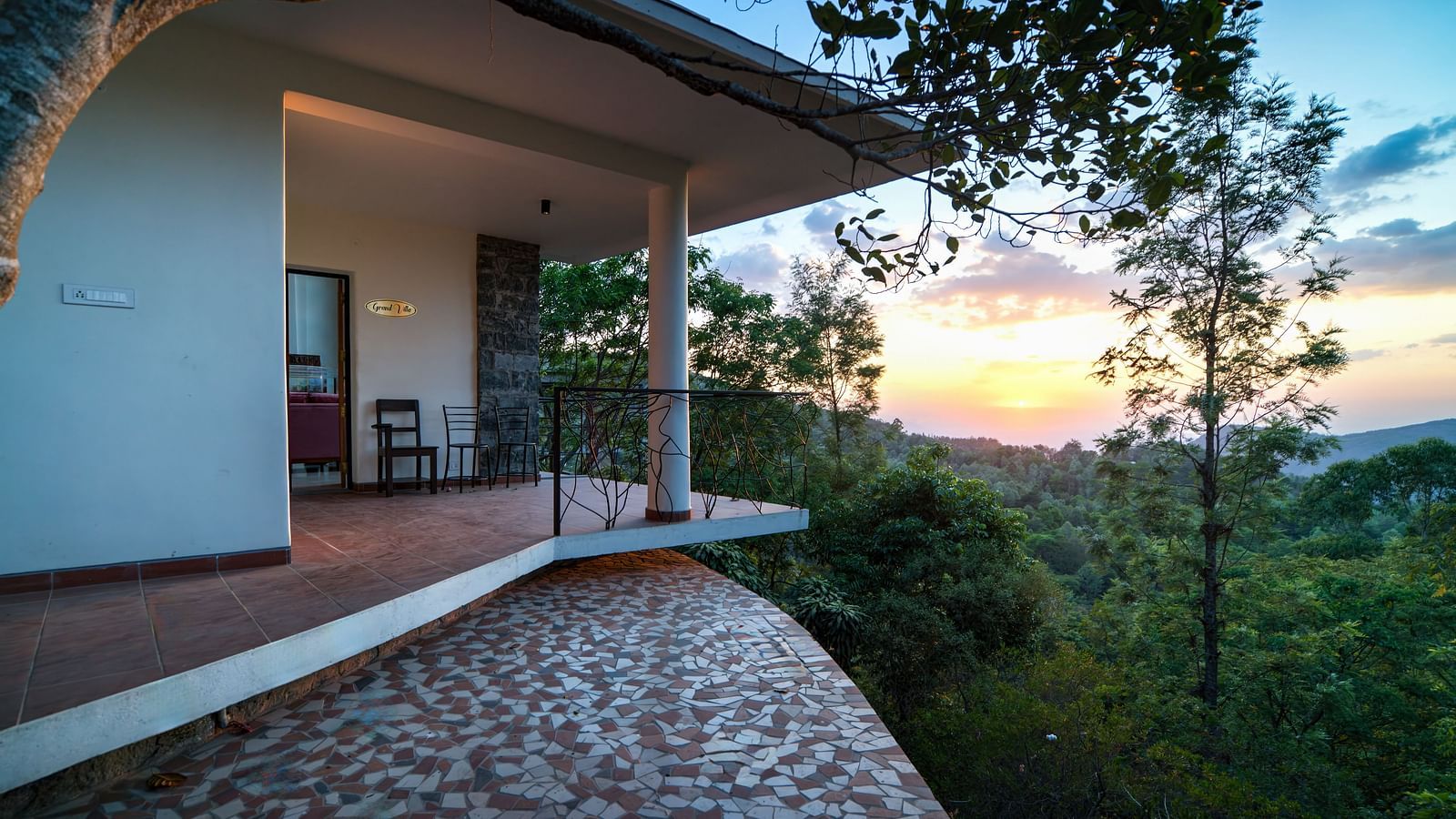 A balcony offering a stunning view of the sunset over a lush green landscape, with a table and chairs for relaxation.  | Mango Hill, Yercaud